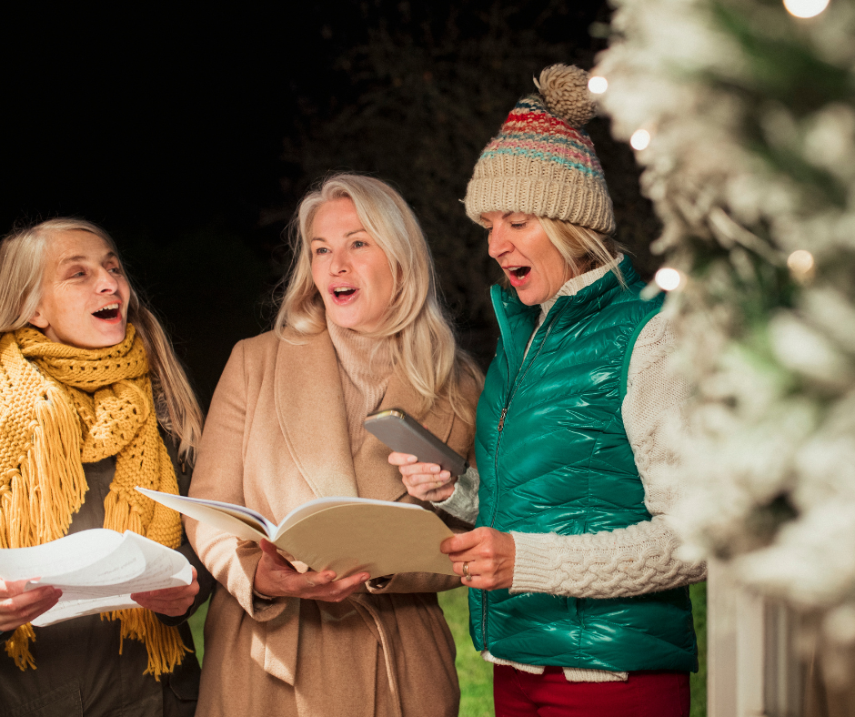 Three women singing carols outdoors at night beside a lit Christmas tree