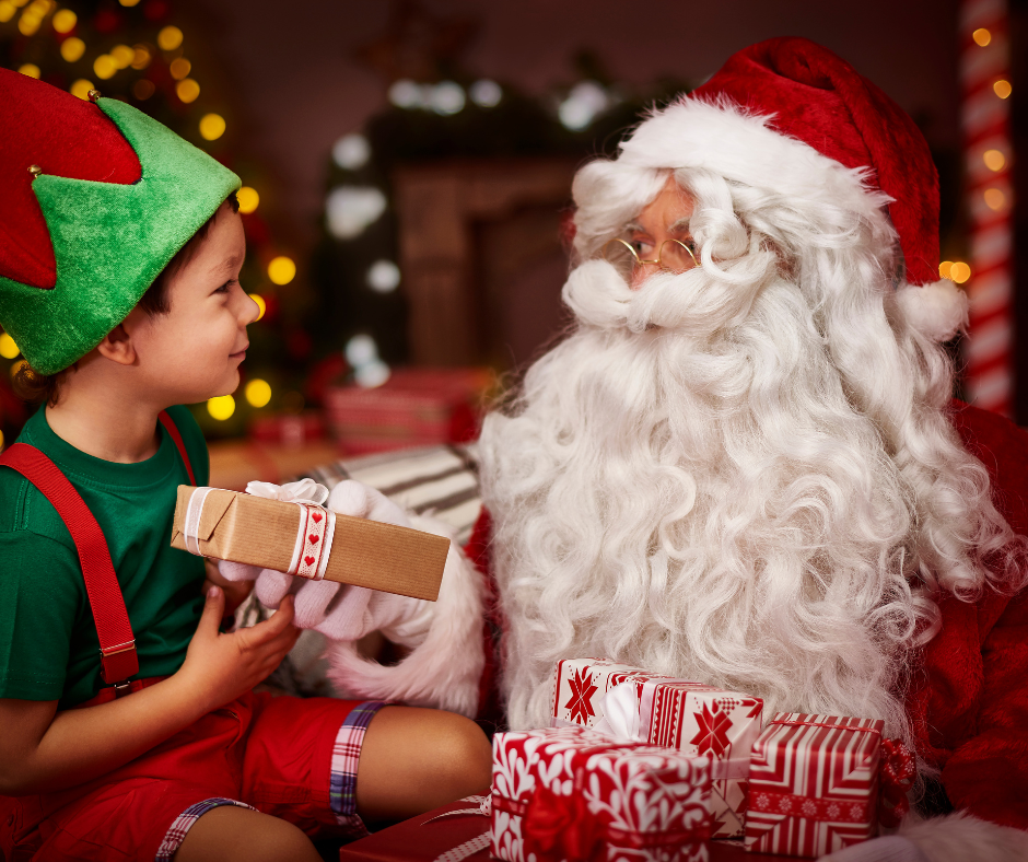 A child in an elf hat handing a wrapped present to Father Christmas