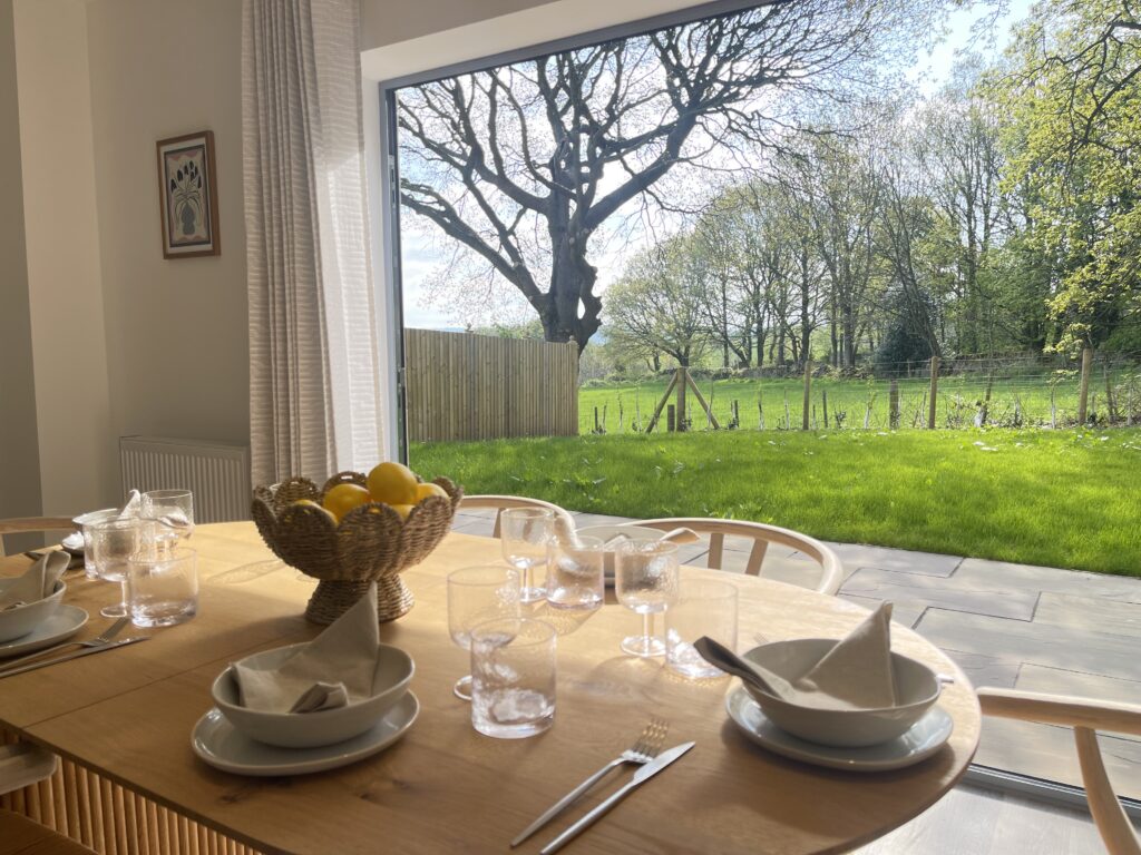 A dining table laid for a meal beside open doors onto a garden and fields